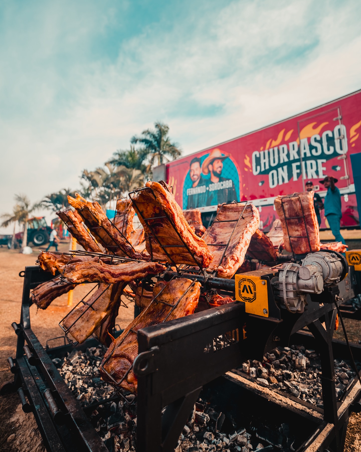 Costelas em churrasqueira girando enquanto assa tendo ao fundo cartas do evento escrito Churrasco on Fire